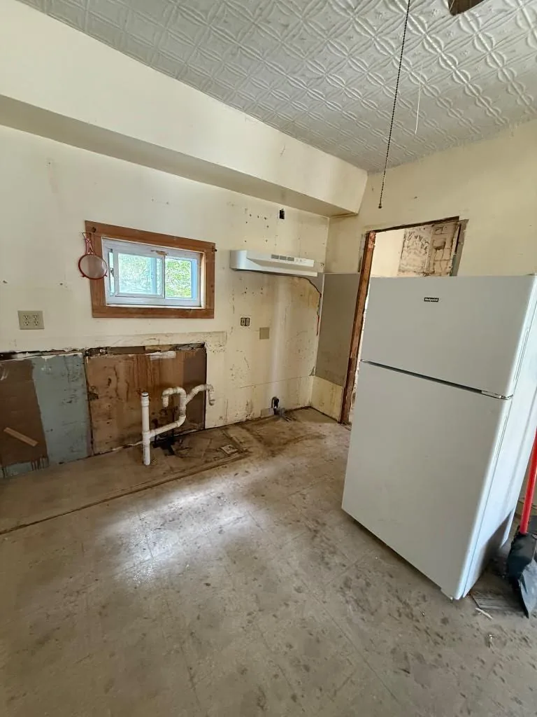 Kitchen before renovation with removed cabinets, exposed plumbing, and refrigerator